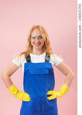 Smiling woman worker wearing protective blue overalls, clear safety goggles, and yellow gloves, standing with hands on hips, ready for a job on a pink background Smiling woman worker wearing protective blue overalls, clear safety goggles, and yellow gloves, standing with hands on hips, ready for a job on a pink background 140835703