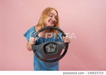 Happy woman with red hair holding a worn car steering wheel on a pink background, expressing joy and freedom related to driving and vehicle ownership Happy woman with red hair holding a worn car steering wheel on a pink background, expressing joy and freedom related to driving and vehicle ownership 140835724