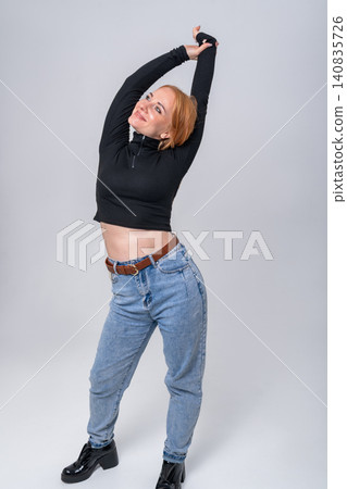 Young woman with red hair stretching her body to the side, expressing happiness and carefree attitude in a casual outfit, standing on a light background 140835726