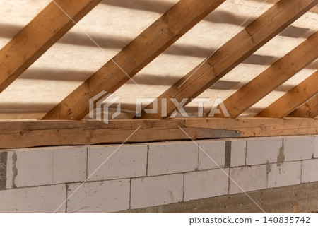 Unfinished house construction showing a close up of wooden roof rafters supporting a white membrane, positioned above a wall built with lightweight concrete blocks Unfinished house construction showing a close up of wooden roof rafters supporting a white membrane, positioned above a wall built with lightweight concrete blocks 140835742