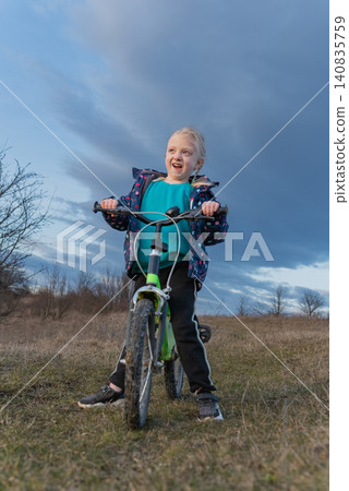 Young girl with blonde braids showing an excited smile while balancing on a bicycle in a grassy field, enjoying outdoor play and childhood adventure under a cloudy sky 140835759