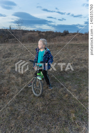 Blonde girl standing confidently with a balance bike in a wide grassy field, looking thoughtfully to the side beneath a cloudy sky, capturing childhood independence and exploration Blonde girl standing confidently with a balance bike in a wide grassy field, looking thoughtfully to the side beneath a cloudy sky, capturing childhood independence and exploration 140835760
