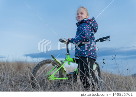 Young blonde girl in a polka dot jacket stands with her bicycle in dry grass, full length, looking away under a wide blue sky, outdoor childhood leisure and freedom Young blonde girl in a polka dot jacket stands with her bicycle in dry grass, full length, looking away under a wide blue sky, outdoor childhood leisure and freedom 140835761