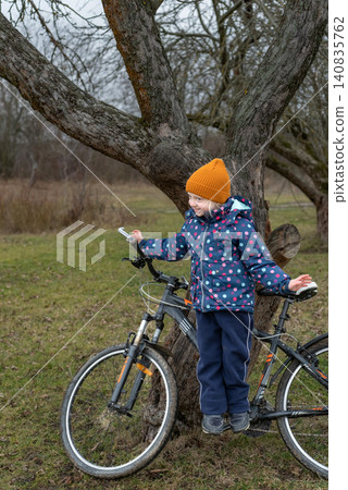 Young girl with orange hat and blue jacket happily standing on a bicycle seat, leaning against a tree trunk in a grassy outdoor setting, exploring and enjoying childhood Young girl with orange hat and blue jacket happily standing on a bicycle seat, leaning against a tree trunk in a grassy outdoor setting, exploring and enjoying childhood 140835762