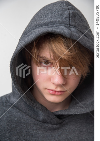 Young person in a gray hooded sweatshirt with hair partly covering their face, looking directly at the camera with a serious, intense expression and contemplative gaze about identity Young person in a gray hooded sweatshirt with hair partly covering their face, looking directly at the camera with a serious, intense expression and contemplative gaze about identity 140835780