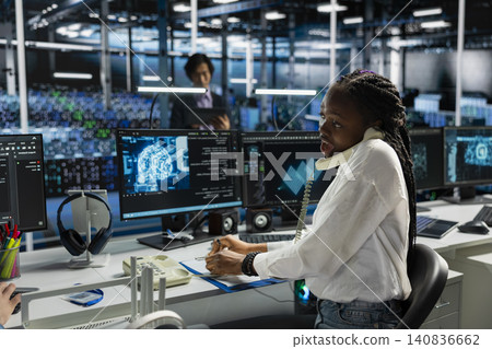 African American woman answers a phone call in a server hub, using an AI assistant to provide technical support to users. African American woman answers a phone call in a server hub, using an AI assistant to provide technical support to users. 140836662