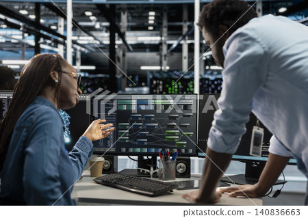 Manager supervises a technician coding in a server farm, overseeing repairs on data center racks used for computational operations. 140836663