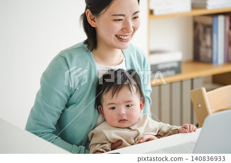 A woman working remotely while holding her zero-year-old baby. A woman working remotely while holding her zero-year-old baby. 140836933