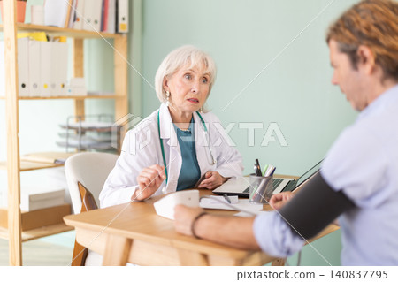 Senior woman doctor measures male patients blood pressure at clinic Senior woman doctor measures male patients blood pressure at clinic 140837795