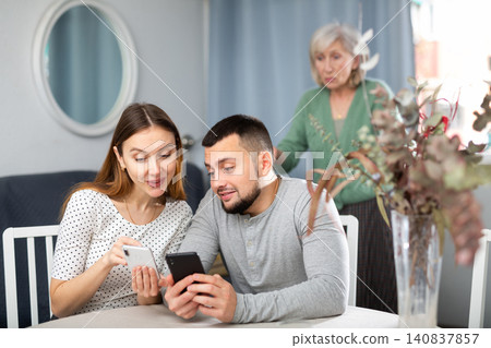 Couple using smartphones at home, displeased senior woman in background Couple using smartphones at home, displeased senior woman in background 140837857