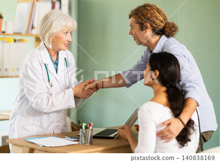 Friendly elderly doctor shaking hands with young couple Friendly elderly doctor shaking hands with young couple 140838169