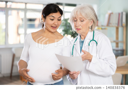 Friendly elderly doctor explaining good diagnostic results to happy pregnant Friendly elderly doctor explaining good diagnostic results to happy pregnant 140838376