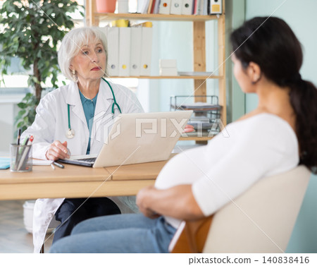 Elderly female doctor talking with pregnant patient at table Elderly female doctor talking with pregnant patient at table 140838416