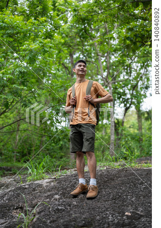 Asian male hiker standing on rocky terrain, admiring the forest during a trek on a mountain trail Asian male hiker standing on rocky terrain, admiring the forest during a trek on a mountain trail 140840892