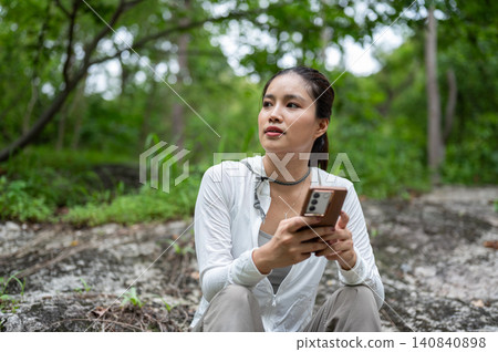Asian female hiker holding smartphone while resting on rocky terrain in forest on a mountain trail 140840898