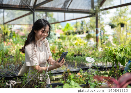 Asian woman visitor taking plant picture with phone while walking through greenhouse nursery garden. Asian woman visitor taking plant picture with phone while walking through greenhouse nursery garden. 140841513