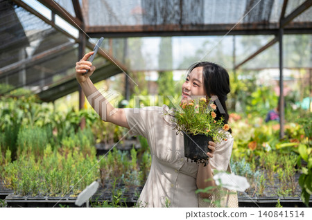 Asian woman visitor taking a selfie with flowering potted plant in greenhouse nursery garden center. Asian woman visitor taking a selfie with flowering potted plant in greenhouse nursery garden center. 140841514