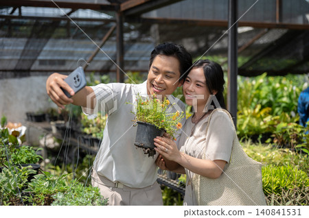 Asian man and woman couple holding a flowering plant taking a selfie in a greenhouse nursery garden. Asian man and woman couple holding a flowering plant taking a selfie in a greenhouse nursery garden. 140841531