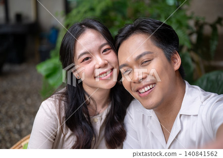 Happy asian man and woman couple leaning together smiling for close up selfie in a green garden cafe Happy asian man and woman couple leaning together smiling for close up selfie in a green garden cafe 140841562