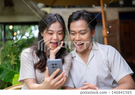 Surprised asian man and woman couple looking at phone together while enjoying time in a garden cafe. Surprised asian man and woman couple looking at phone together while enjoying time in a garden cafe. 140841565