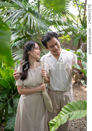 Asian man and woman couple admiring plants and trees while walking through a tropical garden at cafe Asian man and woman couple admiring plants and trees while walking through a tropical garden at cafe 140841576