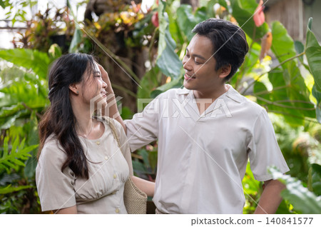 Asian man and woman couple chatting happily while taking a stroll in a tropical garden at green cafe Asian man and woman couple chatting happily while taking a stroll in a tropical garden at green cafe 140841577