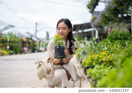 Pretty asian woman holding a potted plant looking closely at flowers while sitting in nursery garden Pretty asian woman holding a potted plant looking closely at flowers while sitting in nursery garden 140841597