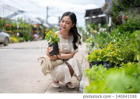 Pretty asian woman admiring potted plant while sitting crouching beside flower bed at nursery garden Pretty asian woman admiring potted plant while sitting crouching beside flower bed at nursery garden 140841598