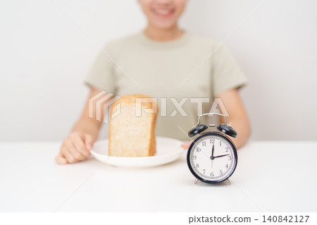 Happy smiling woman with whole wheat bread and alarm clock. concept of intermittent fasting, IF, meal timing, and choosing healthy carbohydrates for a balanced diet and weight loss management Happy smiling woman with whole wheat bread and alarm clock. concept of intermittent fasting, IF, meal timing, and choosing healthy carbohydrates for a balanced diet and weight loss management 140842127