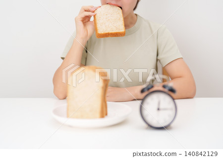 Happy smiling woman eating whole wheat bread with alarm clock. concept of intermittent fasting, IF, meal timing, and choosing healthy carbohydrates for a balanced diet and weight loss management Happy smiling woman eating whole wheat bread with alarm clock. concept of intermittent fasting, IF, meal timing, and choosing healthy carbohydrates for a balanced diet and weight loss management 140842129