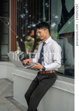 Asian businessman office worker seated outdoors using laptop beside a glass facade of the building. Asian businessman office worker seated outdoors using laptop beside a glass facade of the building. 140842267