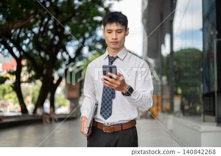 Asian businessman office worker looking at smartphone while carrying laptop walking outside building Asian businessman office worker looking at smartphone while carrying laptop walking outside building 140842268