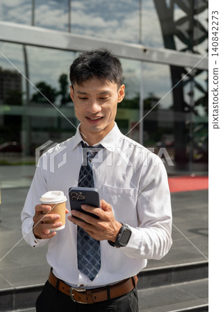 Asian businessman office worker holding coffee looking at smartphone outside building on sunny day. Asian businessman office worker holding coffee looking at smartphone outside building on sunny day. 140842273