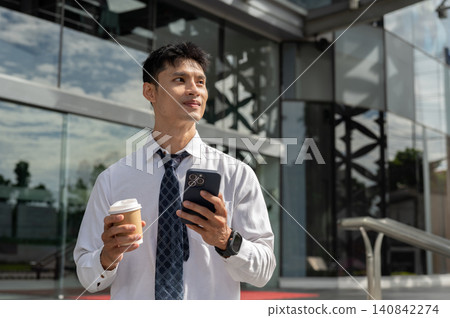 Asian businessman office worker holding coffee cup looking away from phone at a building's entrance. 140842274
