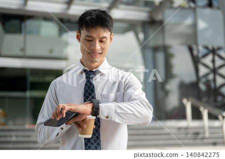 Asian businessman office worker checking time on watch while holding coffee cup outside the building 140842275