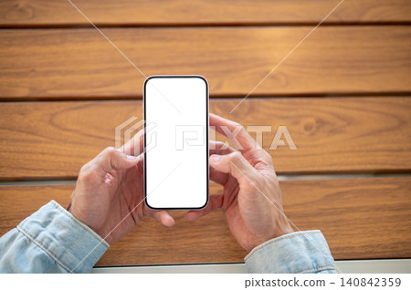 Close up of a man hands holding blank white screen smartphone over wooden table, ideal app template. 140842359