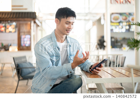 Asian man using a smartphone in cafe environment, enjoying mobile communication and online services. Asian man using a smartphone in cafe environment, enjoying mobile communication and online services. 140842572