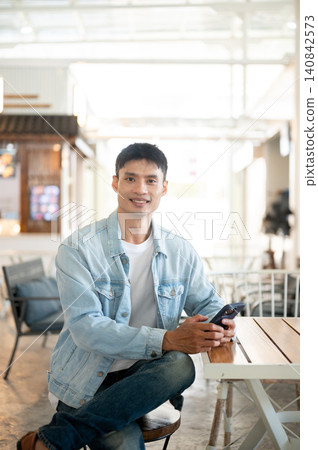 Asian man customer in denim and jeans sitting at table in cafe with smartphone showcasing lifestyle. Asian man customer in denim and jeans sitting at table in cafe with smartphone showcasing lifestyle. 140842573