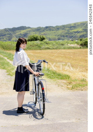 A high school girl pushes her bicycle and looks back in the middle of a rural landscape. A high school girl pushes her bicycle and looks back in the middle of a rural landscape. 140842848