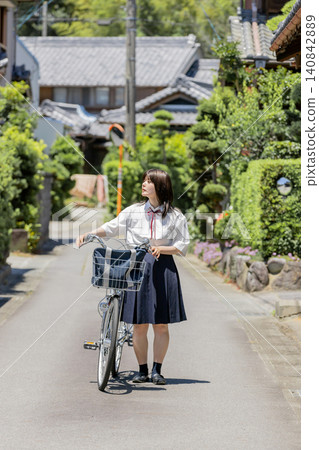 A high school girl walks along a school route in a rural residential area, pushing her bicycle. A high school girl walks along a school route in a rural residential area, pushing her bicycle. 140842889