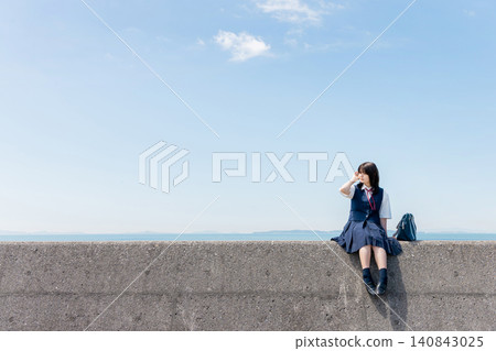 一位高中女生坐在海邊的防波堤上，背景是一片藍天。 140843025