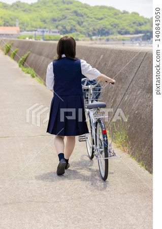 The back view of a high school girl walking along a seaside school route, pushing her bicycle. The back view of a high school girl walking along a seaside school route, pushing her bicycle. 140843050