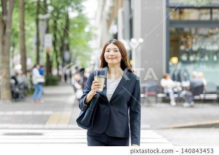 一位面帶微笑的女商人手裡拿著一杯外帶咖啡,走在城市裡。 一位面帶微笑的女商人手裡拿著一杯外帶咖啡,走在城市裡。 140843453