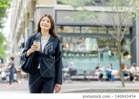一位面帶微笑的女商人手裡拿著一杯外帶咖啡,走在城市裡。 一位面帶微笑的女商人手裡拿著一杯外帶咖啡,走在城市裡。 140843454