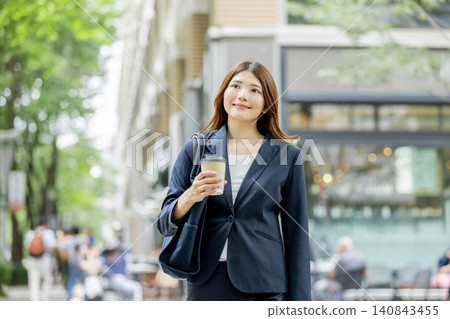 一位面帶微笑的女商人手裡拿著一杯外帶咖啡,走在城市裡。 一位面帶微笑的女商人手裡拿著一杯外帶咖啡,走在城市裡。 140843455