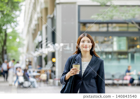 一位面帶微笑的女商人手裡拿著一杯外帶咖啡，走在城市裡。 140843540