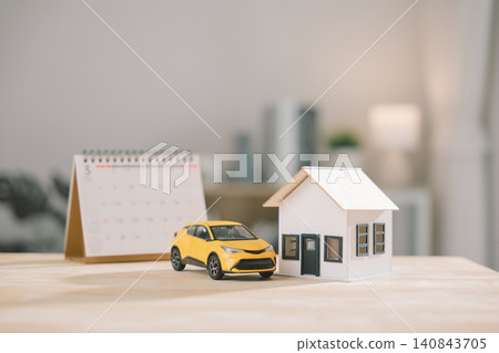 White house model beside yellow car and desk calendar on wooden surface shows real estate investment, while residential rental or vehicle finance and construction insurance protects wealth White house model beside yellow car and desk calendar on wooden surface shows real estate investment, while residential rental or vehicle finance and construction insurance protects wealth 140843705