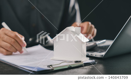 Investor signing mortgage loan interest rate payment real estate model planning property investment documents beside small home miniature using laptop computer at dark office wooden desk Investor signing mortgage loan interest rate payment real estate model planning property investment documents beside small home miniature using laptop computer at dark office wooden desk 140843709