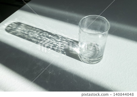 Glass cup casts shadow with light reflections on surface. Transparent object creates pattern of highlights and lines. Concept reflects light, shadow, minimal still life composition Glass cup casts shadow with light reflections on surface. Transparent object creates pattern of highlights and lines. Concept reflects light, shadow, minimal still life composition 140844048