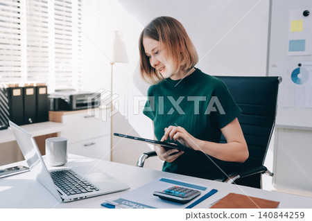 Concept of business working, Businesswoman sitting while writing and checking business analytic document report and holding tablet with computer pc, smartphone on desk in her workstation. Concept of business working, Businesswoman sitting while writing and checking business analytic document report and holding tablet with computer pc, smartphone on desk in her workstation. 140844259